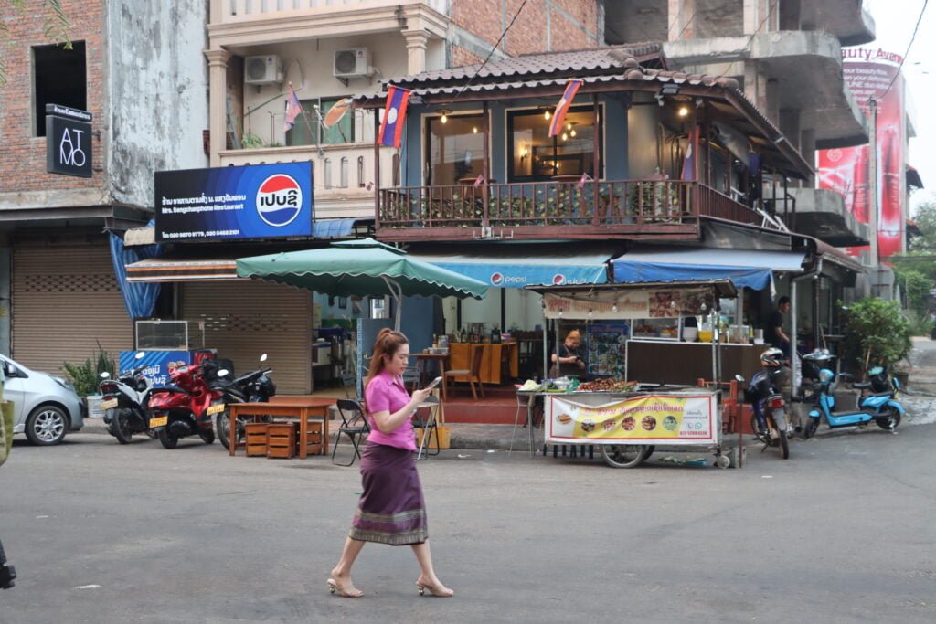 Scène de rue à Vientiane au Laos avec des stands de street food et restaurants. Une femme porte une jupe traditionnelle Sinh et utilise son téléphone portable. Cette photo illustre la fusion entre traditions et modernité.