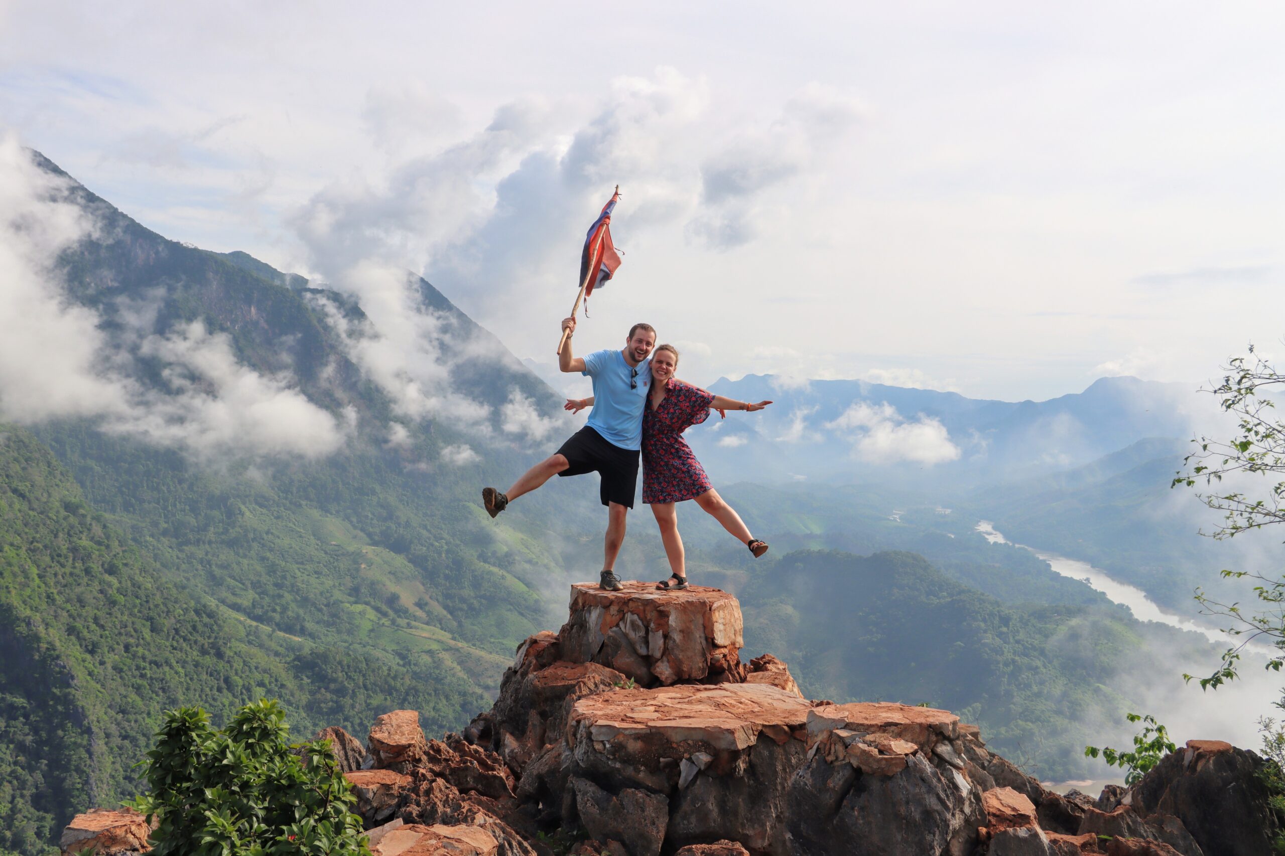 Léo et Cloé, fondateur de l'agence Sourire du Laos célèbrent leur ascension au Laos en brandissant un drapeau sur un sommet montagneux, avec une vue imprenable sur le paysage environnant.